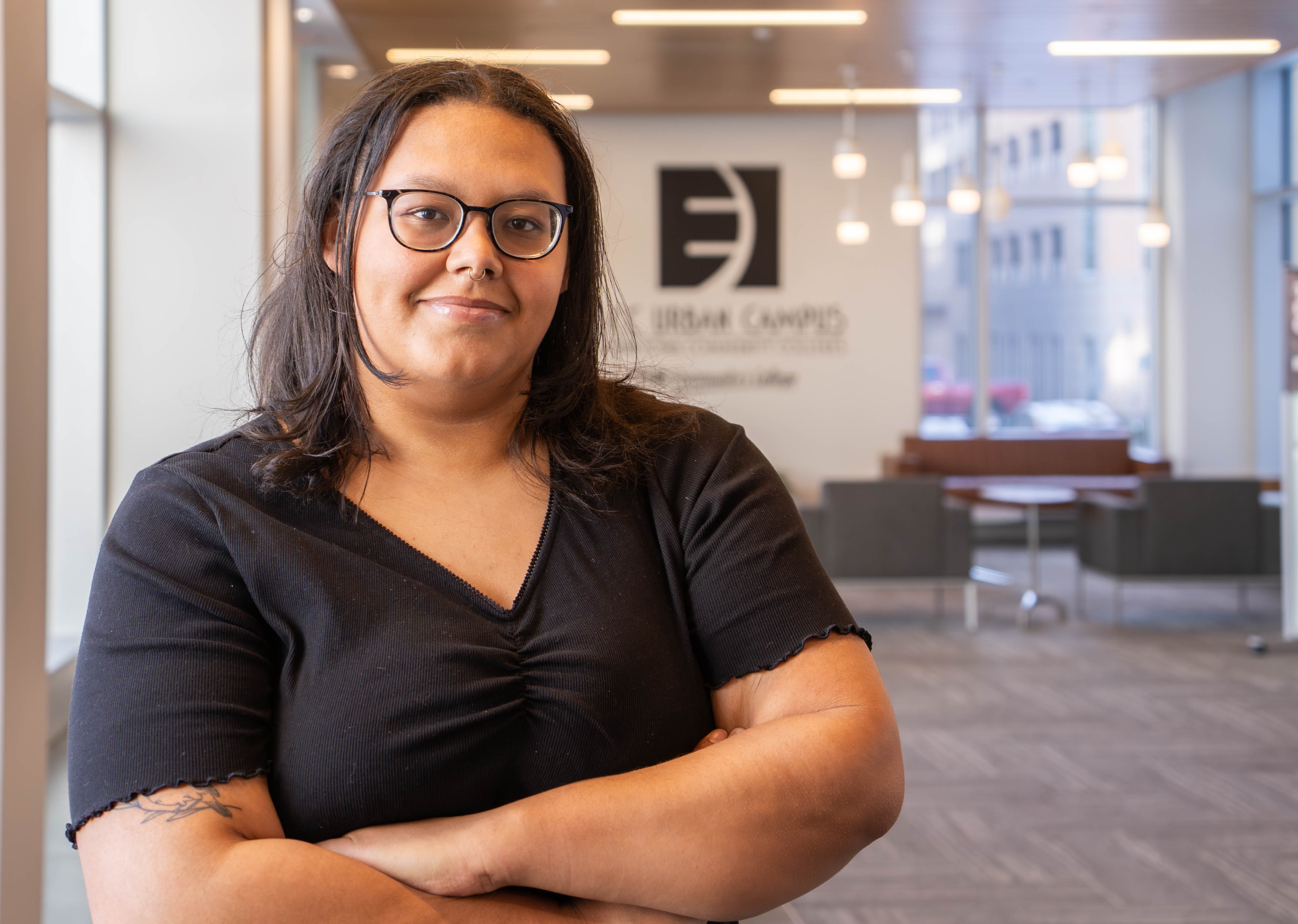 Harli Herrington, EICC alumna, stands indoors with arms crossed, smiling slightly. She wears glasses and a black top, with a softly lit campus lobby and EICC signage in the background.